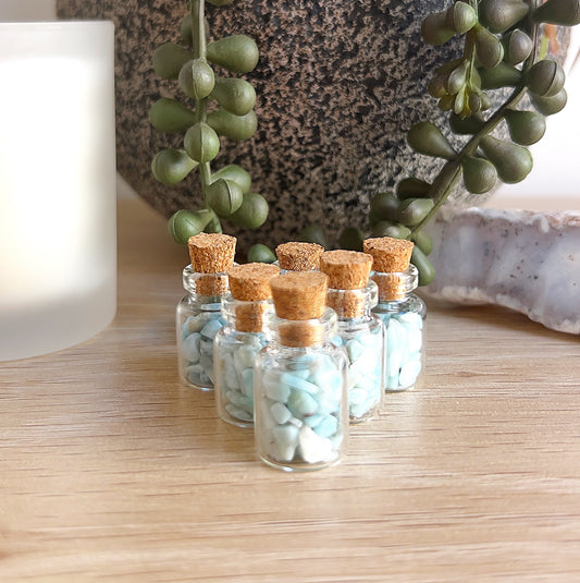 Several small glass bottles filled with soft blue Larimar crystal chips, each sealed with a cork stopper, arranged on a wooden surface beside a succulent plant, white candle, and natural stone, creating a calm and peaceful display. | Simply Esoteric