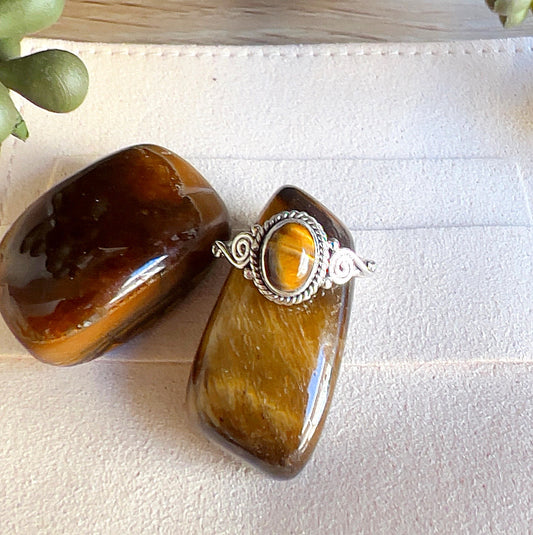 Sterling silver Tiger Eye ring with oval golden-brown stone and decorative swirl detailing, displayed with polished Tiger Eye crystals on a light surface. | Simply Esoteric