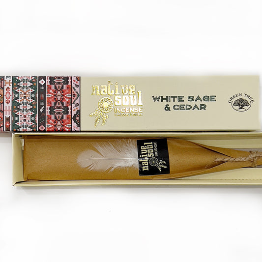 Open box of 'Native Soul Incense Smudge Sticks' in 'White Sage & Cedar' variant, with tribal patterns adorning the left side of the box. The right side has the product name in bold, gold lettering against a cream background, alongside a logo of a tree. Inside, a wrapped smudge stick with a delicate white feather attached, rests on a natural brown paper bed, ready for use in cleansing and aromatic rituals. | Simply Esoteric