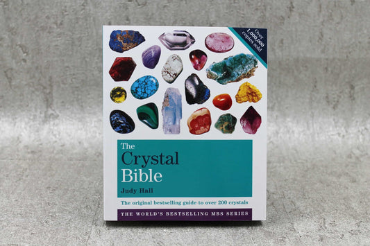 A close-up image of a book titled "The Crystal Bible" by Judy Hall. The cover features a collage of various colorful and textured crystals against a white background, with the title in a bold teal block at the bottom. The book promises an extensive guide to over 200 crystals and includes the tagline "The original bestselling guide to crystals" along with a claim of over one million copies sold, highlighting its popularity and authority on the subject. | Simply Esoteric