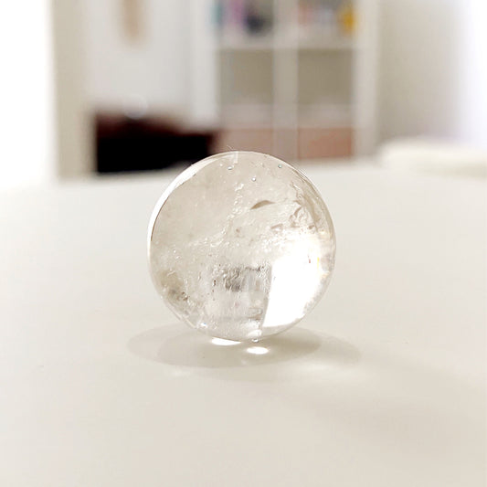 A perfectly polished clear quartz sphere, transparent and gleaming, showcasing natural inclusions and reflecting light beautifully. This sphere is a versatile tool for energy harmonization and spiritual clarity, ideal for meditation and decorative purposes. | Simply Esoteric