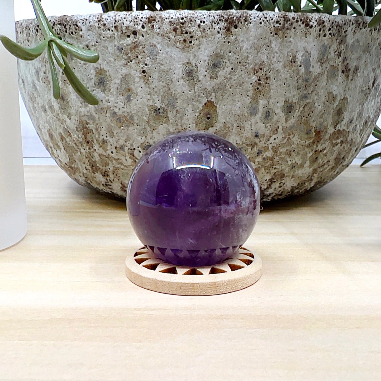 Polished amethyst sphere approximately 3cm in diameter, featuring deep purple tones with natural internal patterns, displayed on a wooden stand against a light wooden surface, with a ceramic pot and white candle in the background. | Simply Esoteric