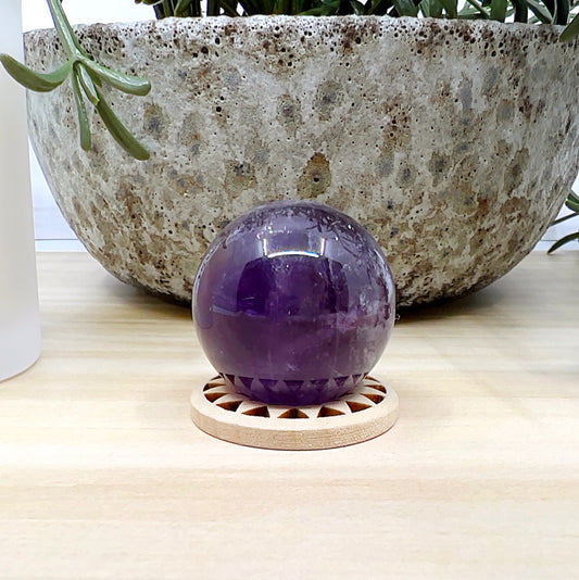 Polished amethyst sphere approximately 3cm in diameter, featuring deep purple tones with natural internal patterns, displayed on a wooden stand against a light wooden surface, with a ceramic pot and white candle in the background. | Simply Esoteric