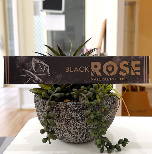 A box of New Moon Aromas Black Rose Natural Incense displayed against a background of greenery. The packaging features an elegant black and gold design with a rose motif, emphasizing its luxurious and natural theme. The box rests on a decorative planter, adding a touch of sophistication to the presentation. | Simply Esoteric