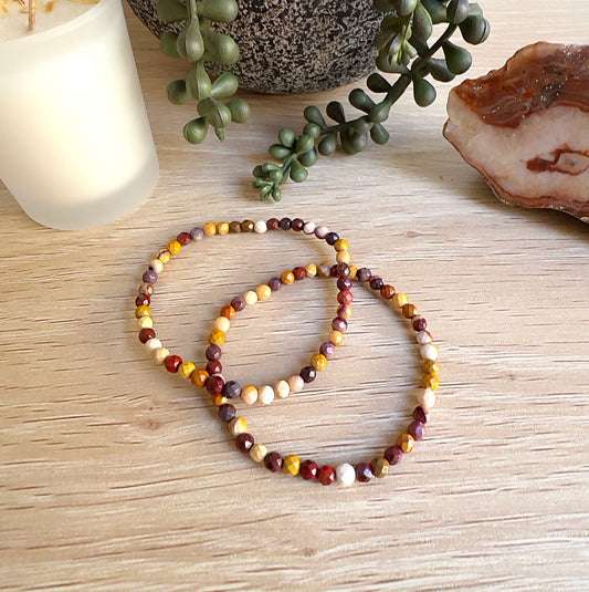 Handmade Mookaite crystal bead bracelets with faceted 4mm beads on stretchy cord, featuring earthy red, yellow, cream, and brown tones, displayed on a wooden surface with a candle, plant, and crystal in the background. | Simply Esoteric