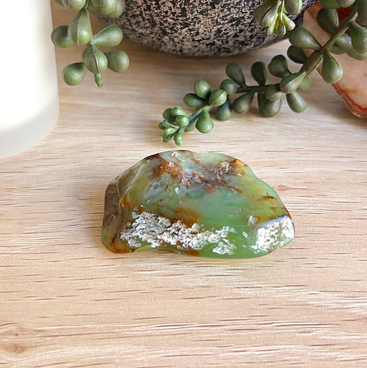 A polished piece of Western Australian chrysoprase, approximately 5cm in size and 43.6g, showcasing vibrant green tones with earthy brown and white mineral inclusions. Displayed on a wooden surface with a succulent plant, candle, and natural crystal slice in the background. | Simply Esoteric