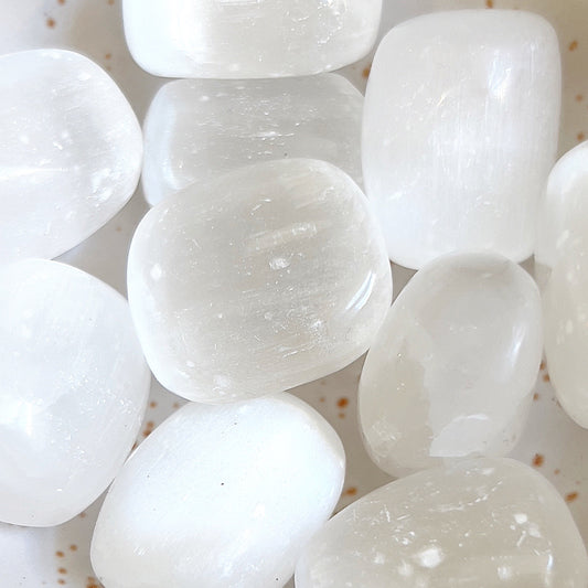 An array of polished selenite tumbled stones with a translucent, icy appearance, nestled together and glinting softly. Their oblong shapes and smooth textures catch and diffuse light, creating a peaceful and cleansing aura in a warm, speckled bowl. | Simply Esoteric