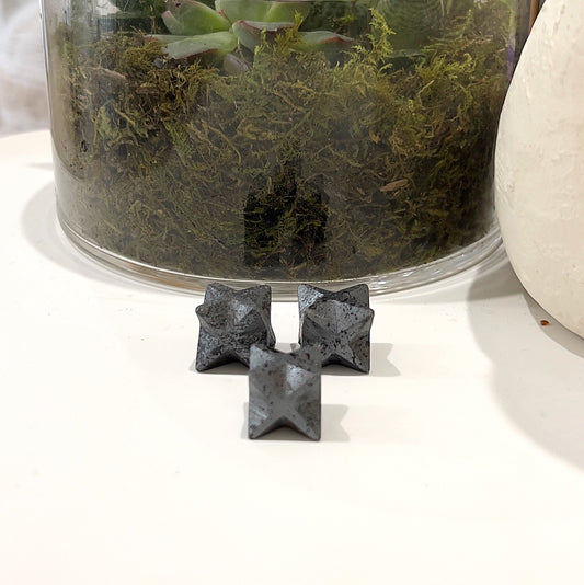 Set of Hematite Merkaba Stars, each approximately 1.5-2cm in size, featuring a dark metallic sheen, displayed with a plant in the background. | Simply Esoteric
