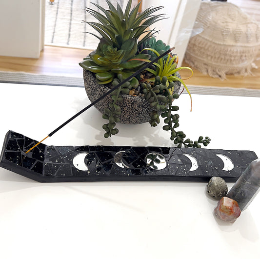 A black mosaic incense burner featuring a design of the moon phases, measuring approximately 5cm wide by 34cm long. The burner rests on a white surface, surrounded by decorative succulents in a textured planter, along with a small cluster of crystals including pyrite, carnelian, and an agate tower. An incense stick is lit, with gentle smoke rising, creating a serene and spiritual ambiance. | Simply Esoteric
