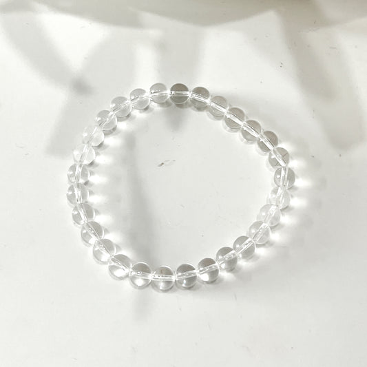 A clear quartz bead bracelet with 6mm round beads strung on a stretchy cord, arranged in a circle on a white background. The beads are transparent and evenly spaced, showcasing their natural clarity and shine. | Simply Esoteric