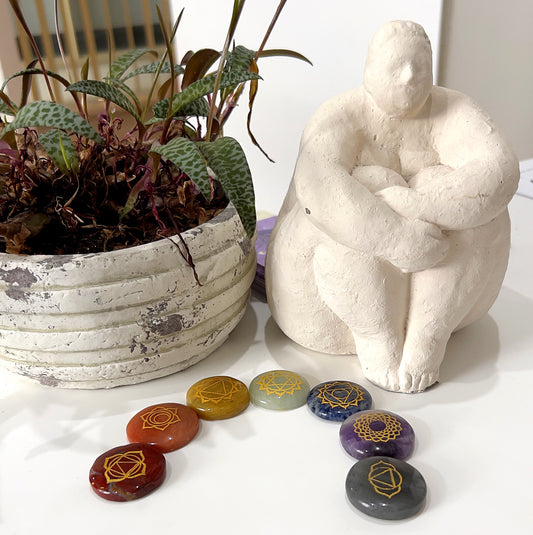 A set of seven polished chakra stones arranged in a line. Each stone is engraved with a unique chakra symbol: Red Jasper for the Root Chakra, Carnelian for the Sacral Chakra, Yellow Aventurine for the Solar Plexus Chakra, Green Aventurine for the Heart Chakra, Sodalite for the Throat Chakra, Agate for the Third Eye Chakra, and Amethyst for the Crown Chakra. The stones are placed on a white surface next to a potted plant and a white sculpture. | Simply Esoteric