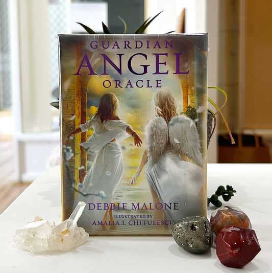 A set of Guardian Angel Oracle cards by Debbie Malone, featuring a 36-card deck with a serene cover illustration of two angelic figures walking through an archway toward a radiant, light-filled background. The deck is accompanied by gemstones, including pyrite and red jasper, placed decoratively beside a quartz crystal cluster. The background includes a green succulent plant in a stone pot, adding a natural and calming aesthetic. | Simply Esoteric