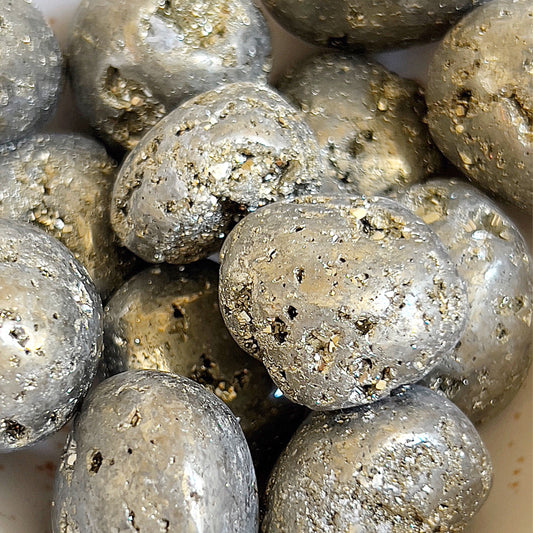 Close-up of pyrite tumbled stones with a lustrous metallic sheen and a rich, gold-like color, their surfaces dotted with natural, crystalline textures. These stones are clustered together, showcasing the raw beauty and glittering facets characteristic of pyrite. | Simply Esoteric