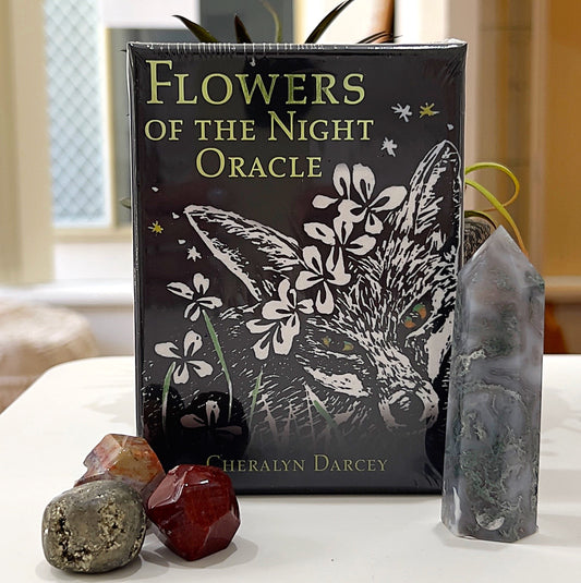 A close-up image of the *Flowers of the Night Oracle* card deck by Cheralyn Darcey. The packaging features an elegant black design with a fox illustration surrounded by white flowers, evoking a mystical, nighttime theme. The deck is displayed upright, accompanied by crystal accents, including red and silver gemstones, and a gray tower crystal in the background. This setup highlights the deck's nature-inspired and spiritual essence. | Simply Esoteric