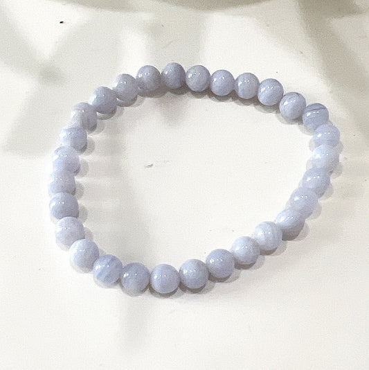 A Blue Lace Agate bead bracelet with 6mm beads strung on a stretchy cord. The bracelet features light blue beads with delicate white bands, arranged in a circular pattern. The background includes a neutral setting with soft lighting, highlighting the serene and calming appearance of the Blue Lace Agate stones. | Simply Esoteric