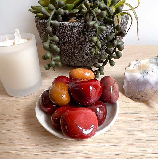 A small white dish filled with polished Mookaite tumbled stones in rich shades of red, yellow, and ochre, displayed on a wooden surface with a candle, plant, and crystal cluster in the background. | Simply Esoteric