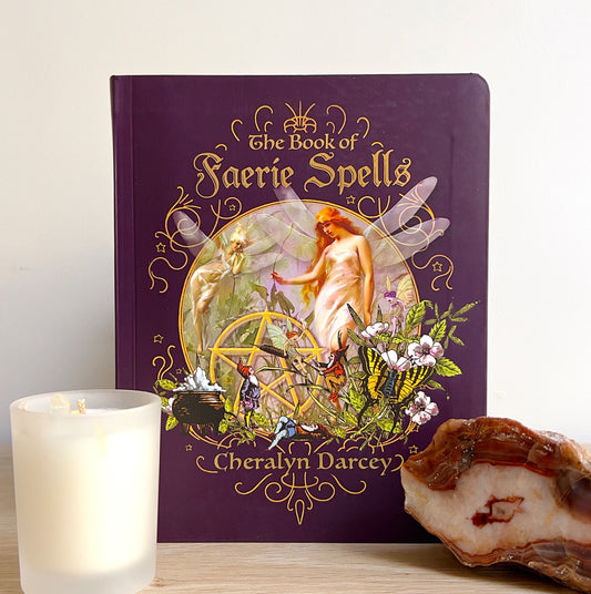 A beautifully bound copy of The Book of Faerie Spells by Cheralyn Darcey, featuring ornate gold detailing and artwork of faeries and nature on a rich purple cover, displayed on a wooden surface with a candle and crystal beside it. | Simply Esoteric