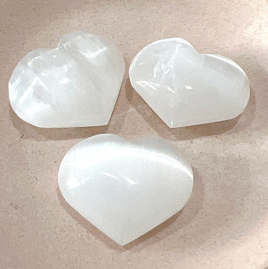Three polished Selenite hearts, approximately 4cm in size, displaying their smooth, translucent surface on a soft pink background.