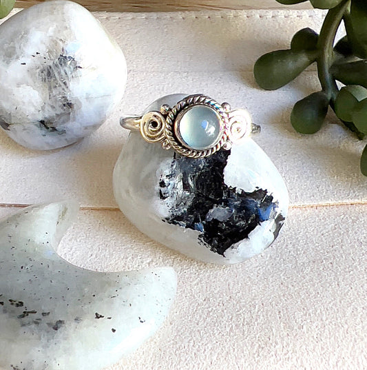 Sterling silver Moonstone ring with a round luminous stone and decorative swirl detailing, displayed with polished Moonstone crystals on a light surface. | Simply Esoteric