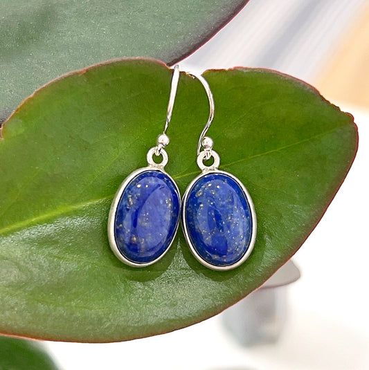 A close-up image of elegant Lapis Lazuli Sterling Silver Drop Earrings resting on a green leaf. The earrings feature deep blue oval-shaped lapis lazuli gemstones with natural golden pyrite flecks, set in a polished sterling silver frame. The hook design adds a delicate and timeless touch to the jewelry. The background highlights the natural and earthy aesthetic. | Simply Esoteric