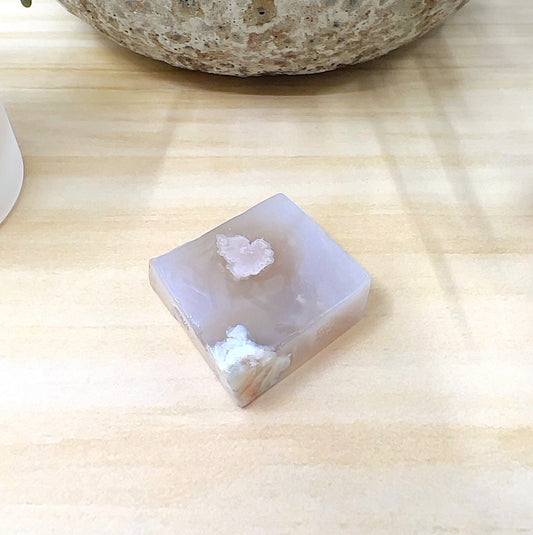 Polished flower agate freeform stone measuring 32mm x 30mm and 13mm thick, featuring soft pinkish-beige tones with white blossom-like inclusions, displayed on a light wooden surface with part of a ceramic pot in the background. | Simply Esoteric