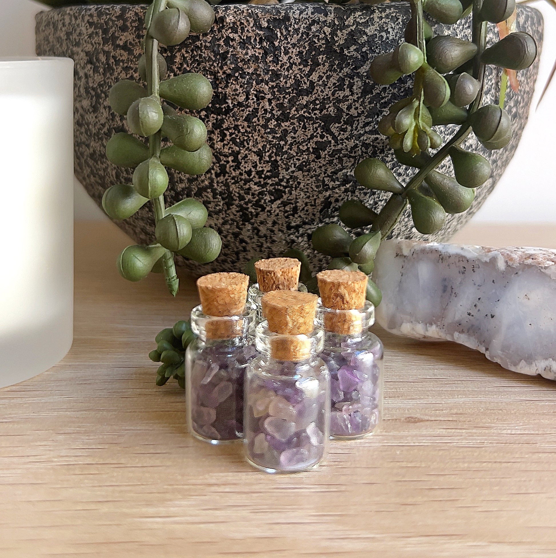 Four small glass bottles filled with delicate amethyst crystal chips, each sealed with a cork stopper, arranged neatly on a wooden surface alongside a succulent plant, white candle, and natural crystal, creating a tranquil and inviting setting. | Simply Esoteric