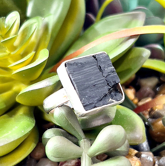 A sterling silver ring featuring a raw black tourmaline stone with a square-cut design, set against a backdrop of lush green succulents and earthy tones. The natural texture of the tourmaline contrasts beautifully with the polished silver band, creating an elegant yet grounding aesthetic. Perfectly suited for showcasing the ring's raw and protective qualities. | Simply Esoteric