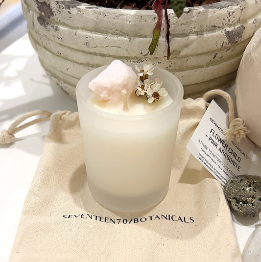A crystal-infused soy wax candle from Seventeen70 Botanicals labeled "Flower Child + Pink Aragonite" sits on a white surface. The candle features a small pink aragonite crystal and scattered white flowers on top, creating a delicate and natural appearance. Accompanied by a label and a burlap drawstring bag, this product emphasizes a connection to nature with its eco-friendly and calming design. | Simply Esoteric