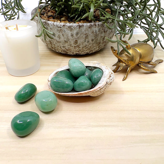 Polished green aventurine tumbled stones with smooth, glossy surfaces, displayed in a shell dish with some stones scattered on a light wooden surface, alongside a white candle, ceramic pot with greenery, and a small brass beetle ornament. | Simply Esoteric