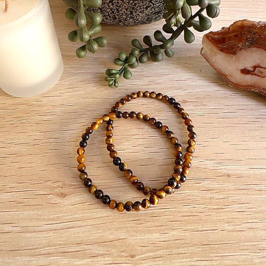 Two Yellow Tiger Eye crystal bead bracelets with approximately 4mm round beads on stretchy cord, displayed on a wooden surface with a candle, greenery, and a polished stone in the background. | Simply Esoteric