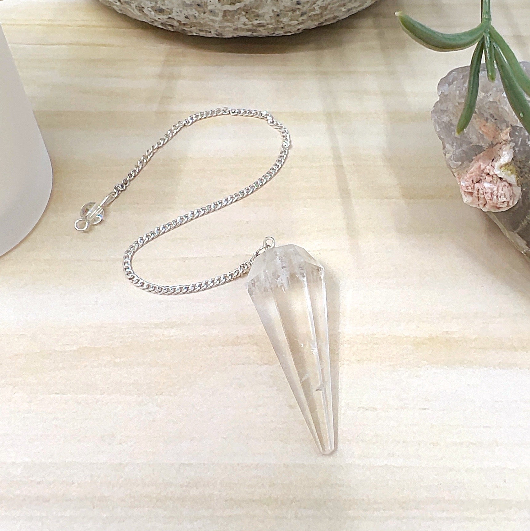 Faceted clear quartz pendulum measuring 4cm x 1.5cm with a metal link chain and clear quartz bead detail, displayed on a light wooden surface with a white candle, crystal cluster, and ceramic pot in the background. | Simply Esoteric