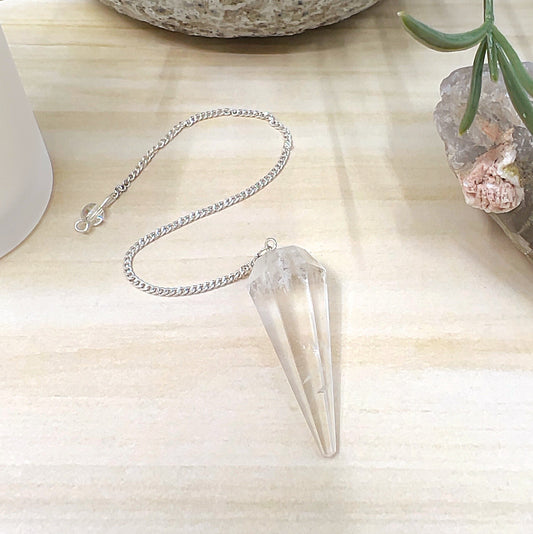 Faceted clear quartz pendulum measuring 4cm x 1.5cm with a metal link chain and clear quartz bead detail, displayed on a light wooden surface with a white candle, crystal cluster, and ceramic pot in the background. | Simply Esoteric