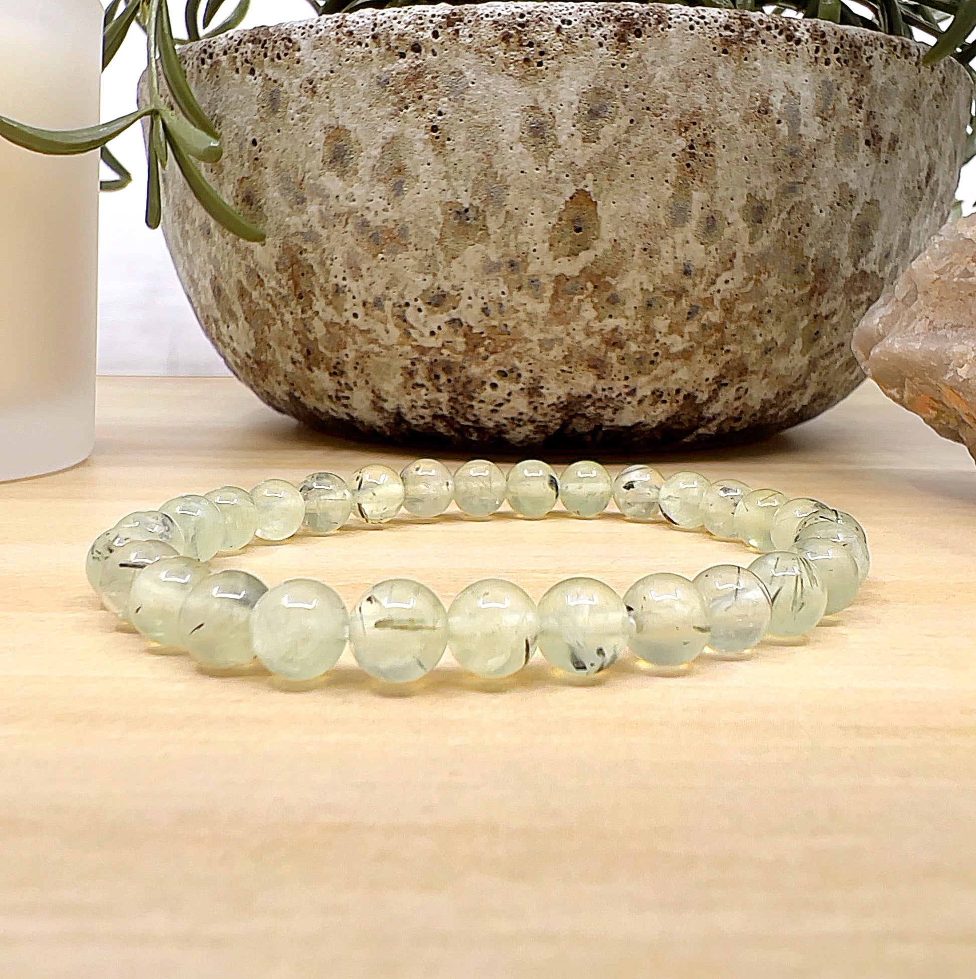 Handmade Prehnite crystal bead bracelet with 6mm round beads on a stretchy cord, featuring soft green tones and natural inclusions, displayed on a light wooden surface with a ceramic pot, candle, and crystal in the background. | Simply Esoteric