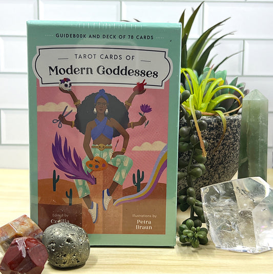 A vibrant box of Tarot Cards of the Modern Goddess by Cecilia Lattari, featuring a colorful illustration of a modern goddess holding various symbols of empowerment, surrounded by a desert landscape with cacti and a rainbow. The set includes a 78-card deck and a guidebook. The box is displayed on a wooden surface with decorative gemstones and crystals placed nearby, adding to the spiritual and empowering theme. | Simply Esoteric